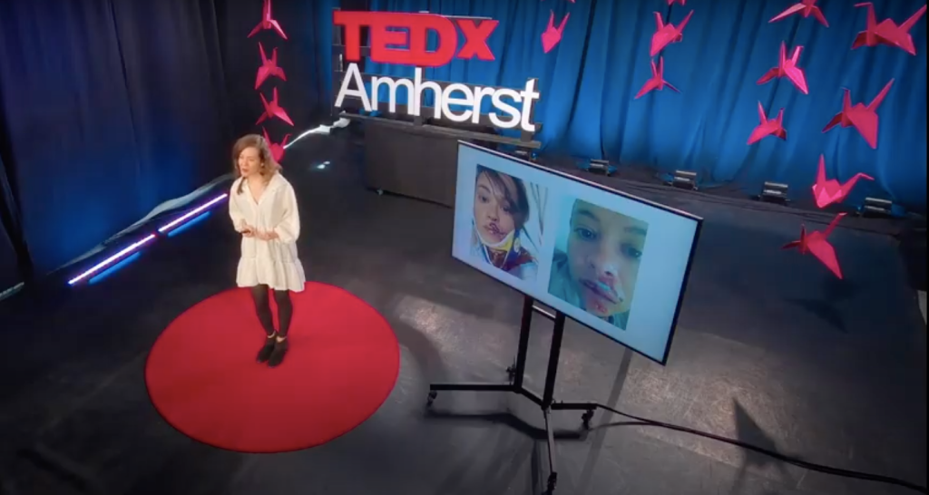 Maria, a short white woman with black hair in a braid, stands on a red circle carpet in a white dress and black shoes. She is in front of a screen with images of her broken face. There are red paper cranes hanging around her, and a sign that says: TEDx Amherst. 